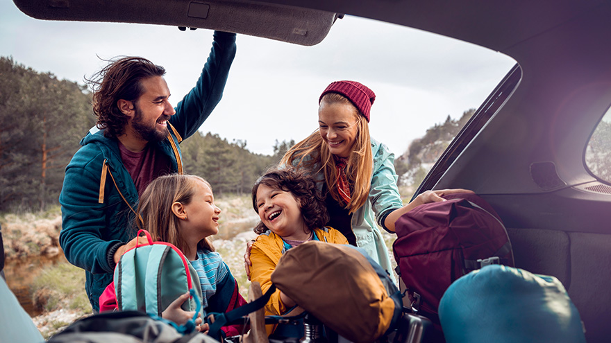 Mother and father standing at open trunk of car with 2 kids getting out hiking gear